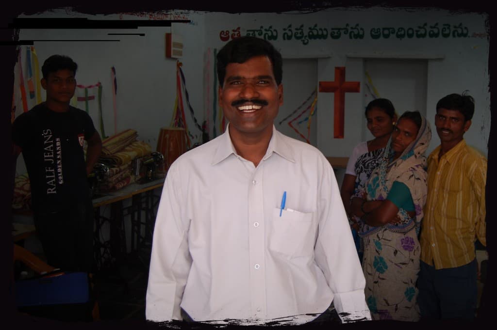 A pastor standing in a church hall with congregation members and vocational sewing machines behind him